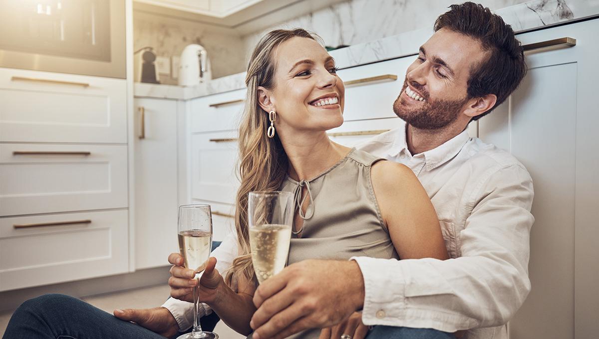 Article Cards Featured Image Celebrate, love and couple with champagne in kitchen for honeymoon, anniversary and valentines day. Happy, relax and young man and woman drinking alcohol on the floor with conversation in marriage